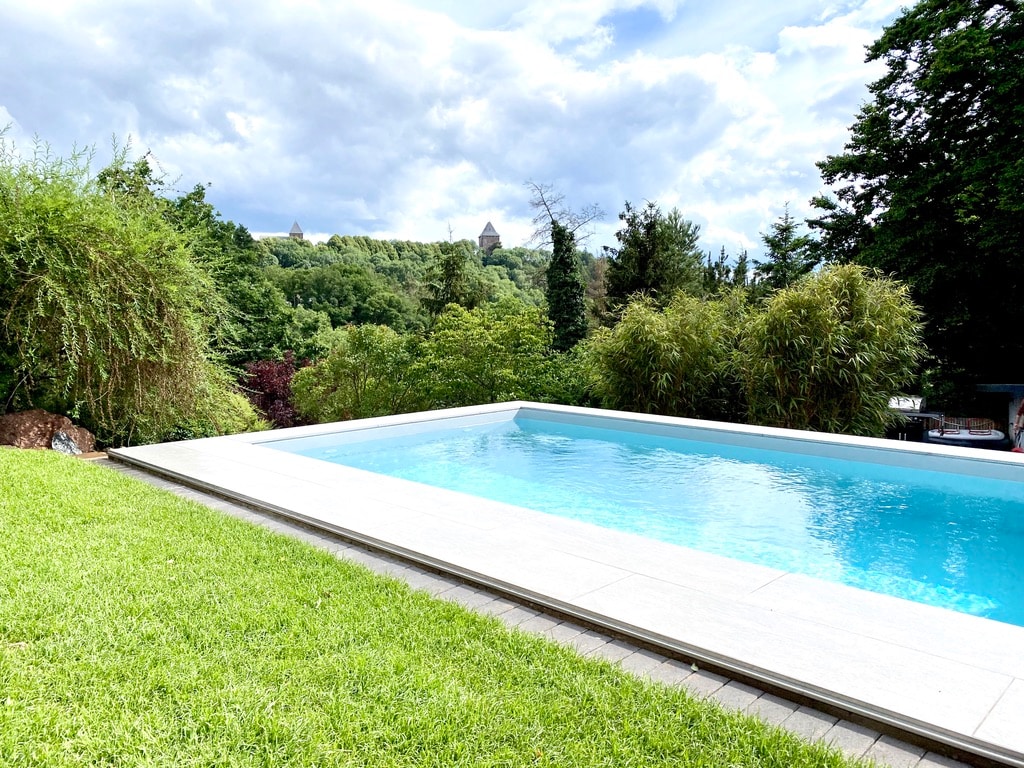 The outdoor pool, surrounded by lush greenery, reflects the sky's varying shades. In the background, a glimpse of a castle is visible among the treetops, creating a peaceful backdrop. The area features well-maintained grass and a clean, modern poolside design.