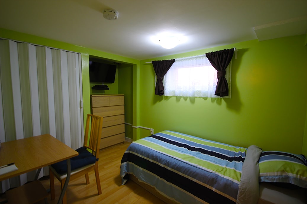 The room features cheerful green walls and a comfortable single bed with striped bedding. A desk and chair are positioned beside the bed, while a wall-mounted TV is visible in the corner. Natural light enters through a window adorned with dark curtains.