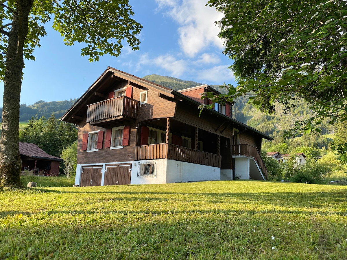 An inviting chalet stands amidst lush greenery, featuring wooden siding and multiple balconies overlooking the expansive lawn. The surrounding landscape includes rolling hills and distant mountains, creating a serene backdrop for outdoor activities.