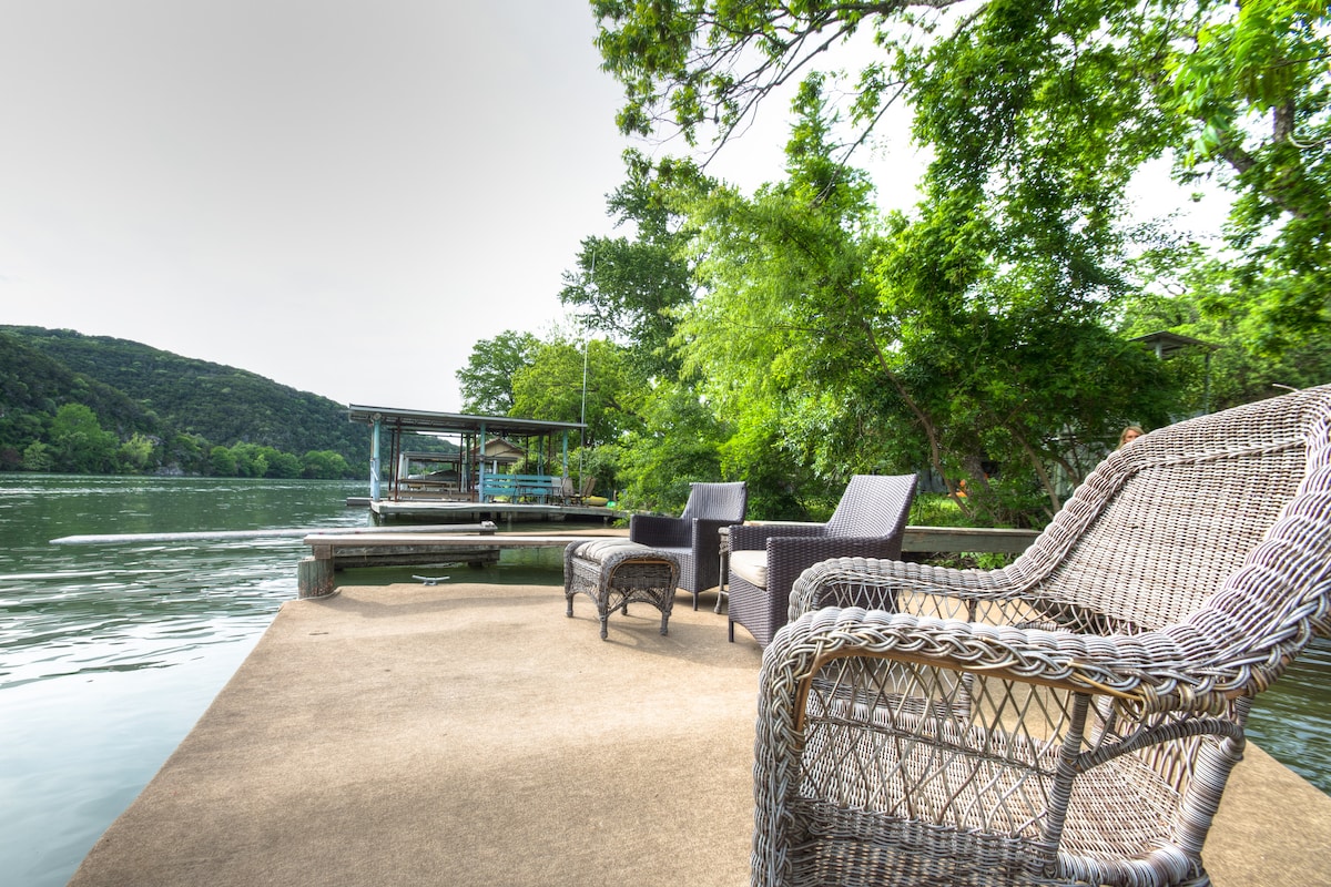 A serene outdoor seating area is situated by the water's edge, featuring a pair of woven chairs and a small side table. Lush greenery surrounds the space, providing a natural backdrop, while a dock is visible in the distance, enhancing the tranquil lakeside setting.