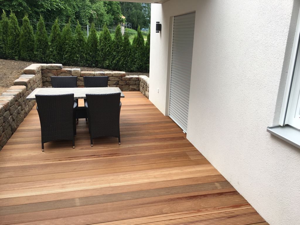 A wooden terrace is furnished with a table and four chairs, providing a comfortable outdoor dining space. Surrounding greenery offers a natural backdrop, while a wall provides additional privacy. The terrace flooring features horizontal wood planks that create a warm, inviting surface.