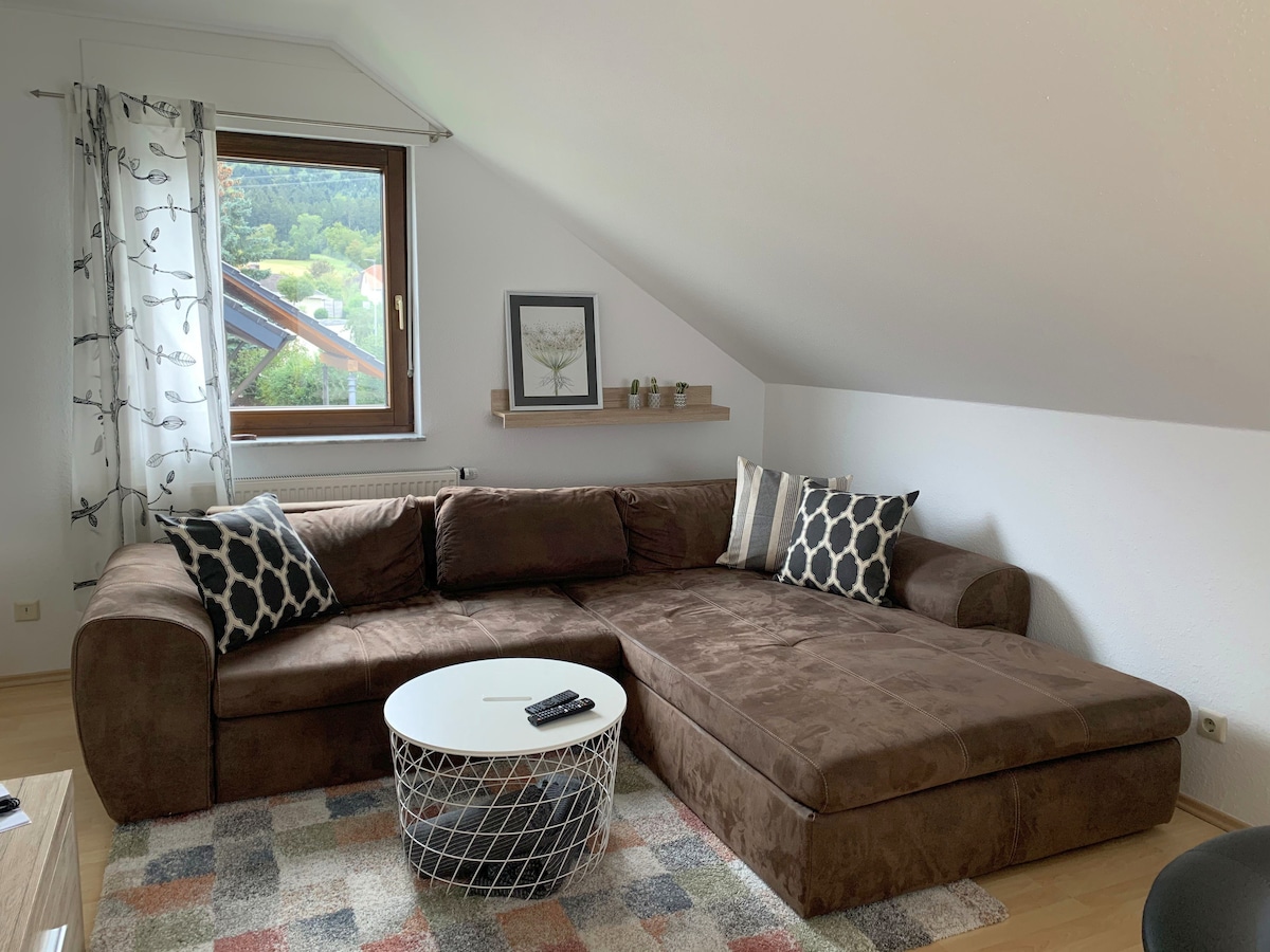 A spacious corner sofa with a cozy, brown upholstery is positioned beneath a slanted ceiling. A round coffee table sits in front, adorned with a remote control. Decorative throw pillows in various patterns add subtle detail to the seating area. A window, partially draped with sheer curtains, allows natural light to fill the space.