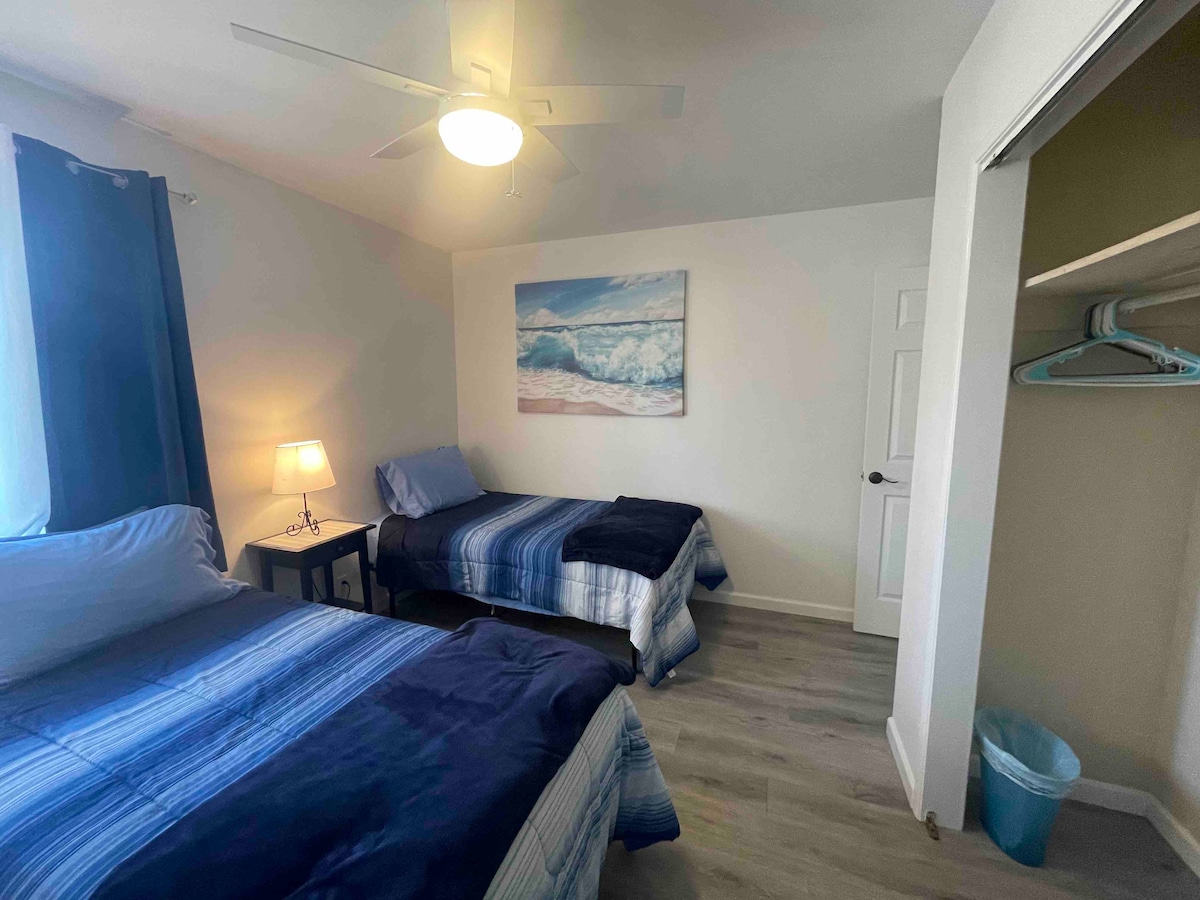 A well-lit bedroom contains two beds, one larger and one twin-sized, both dressed in blue and white striped bedding. A lamp on a nightstand provides additional light. A painting of ocean waves adorns the wall, and a ceiling fan circulates air. A closet is partially visible.