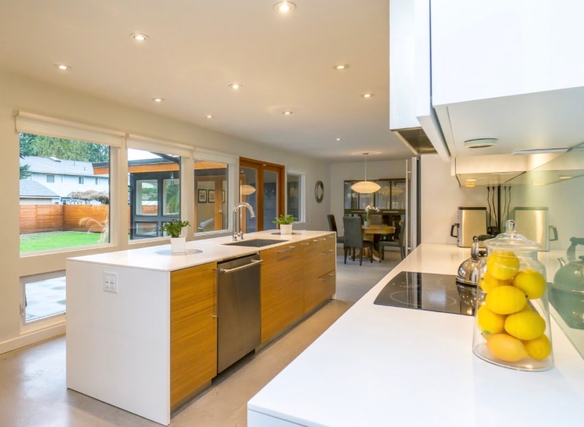 A modern kitchen is featured with a central island that includes a double sink and a stovetop. Natural light floods the space through large windows, highlighting the sleek cabinetry and open layout that connects to a dining area. A bowl of lemons sits on the counter.