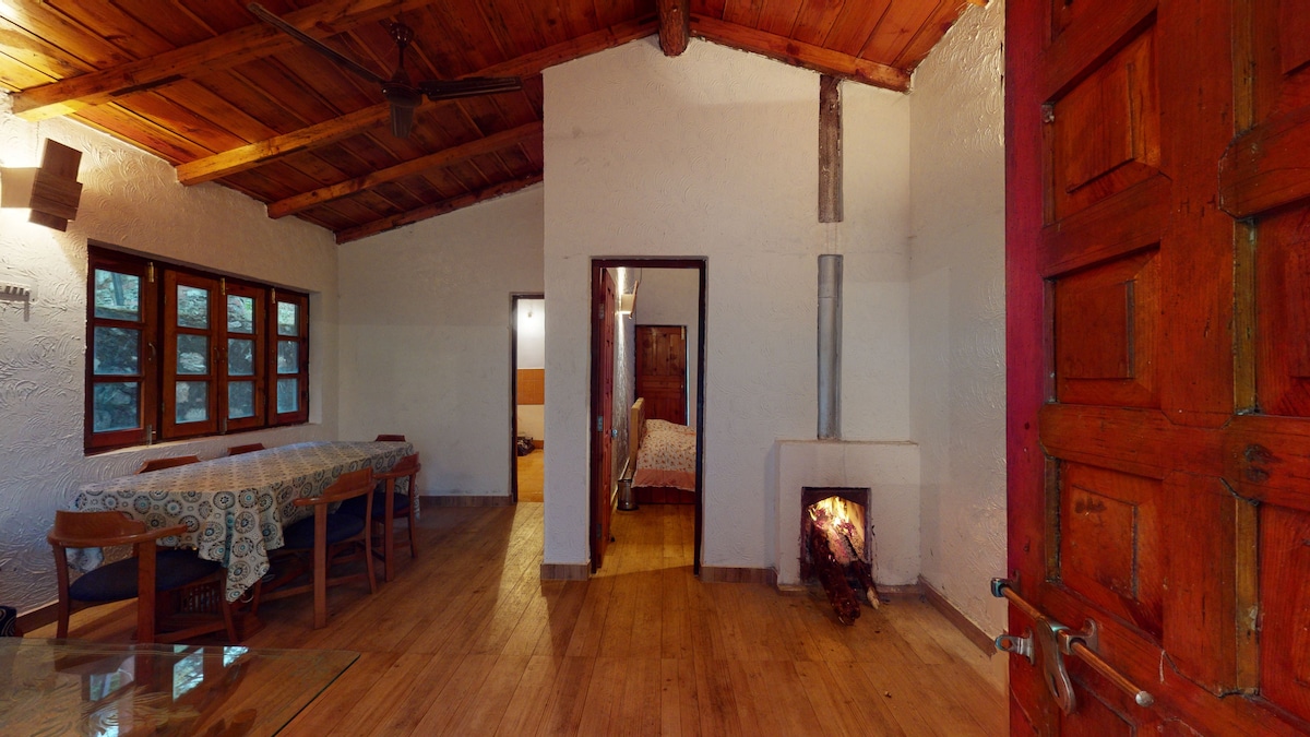 The interior space features wooden beams and a warm wooden floor. A dining area with a patterned tablecloth occupies one side, while a cozy sleeping area is visible in the background, separated by an open doorway. A small stove is located near the entrance.