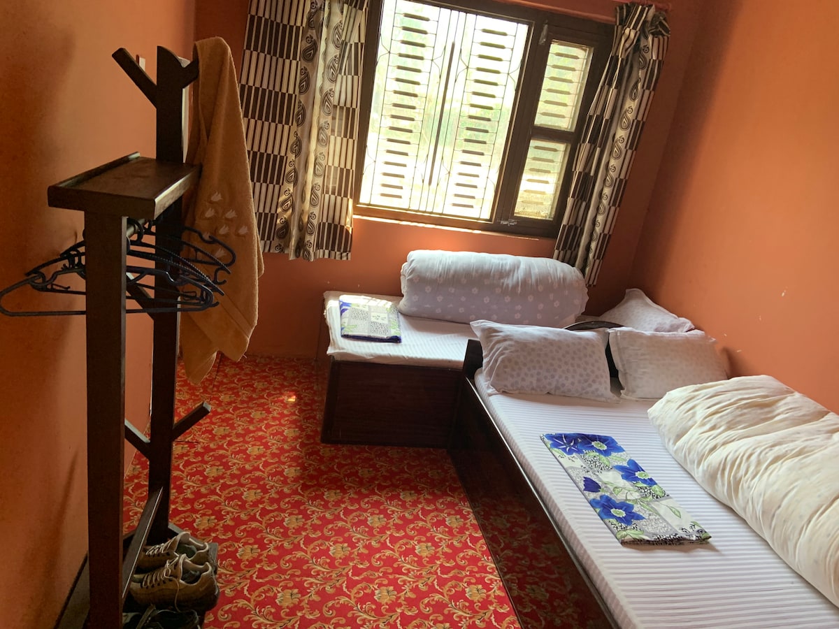 A room is highlighted by two beds, one single and one double, positioned against a vibrant orange wall. Natural light filters through windows with wooden shutters, illuminating the patterned carpet. A wooden rack holds clothes hangers and nearby, a folded towel is neatly arranged.