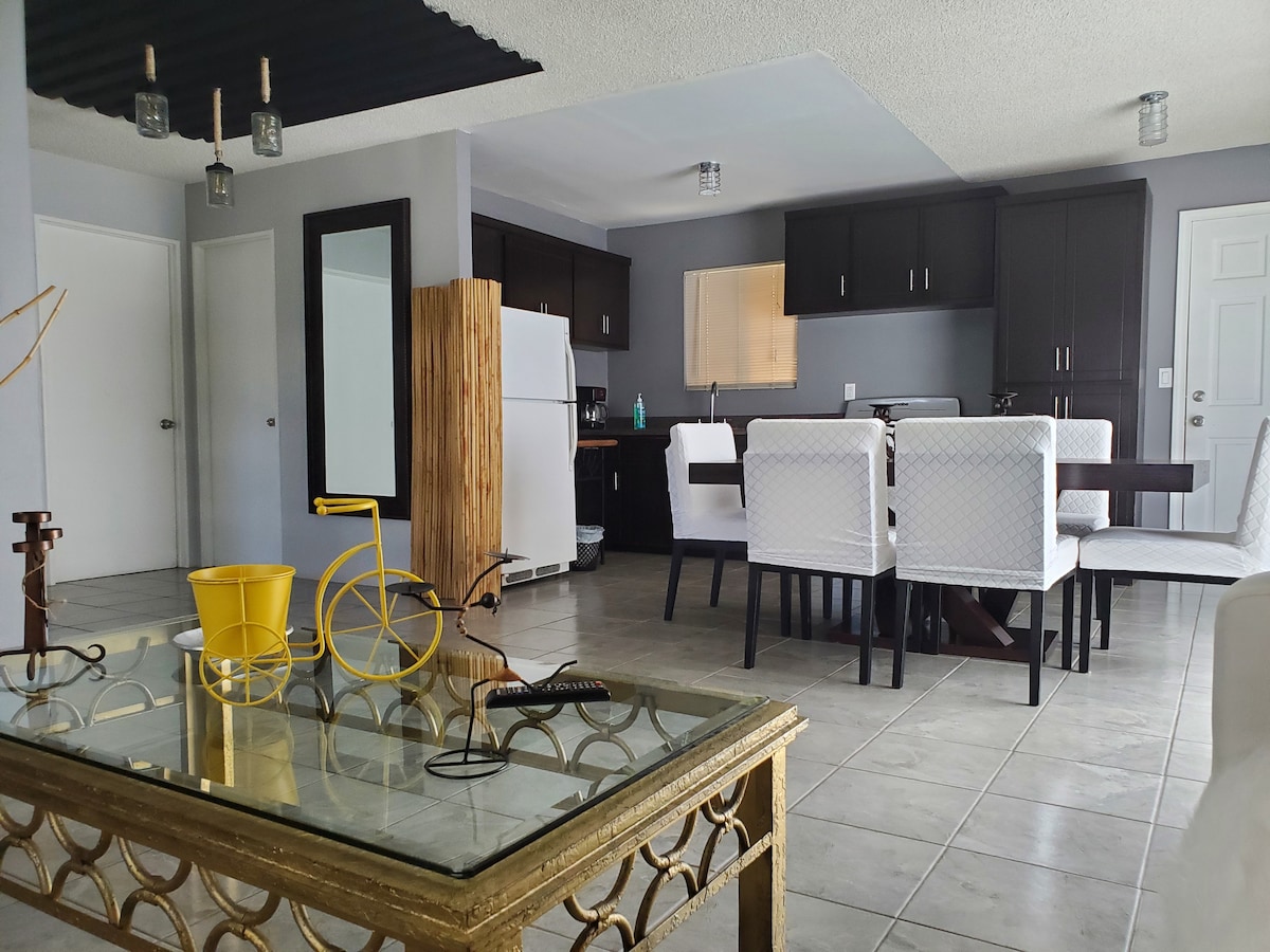 An open living space features a dining area with white chairs around a dark wooden table. A modern kitchen is visible in the background, equipped with sleek cabinets and a refrigerator. A glass coffee table with decorative items is centrally located.
