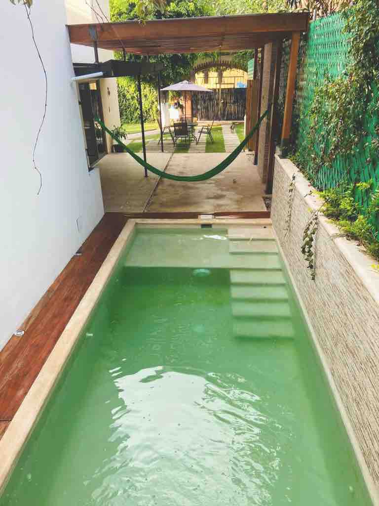 An inviting pool is featured, surrounded by wooden decking and a stone wall. A hammock hangs nearby, alongside a relaxing area with lounge chairs under a shaded umbrella, and a landscaped green wall adds to the tranquil outdoor space.