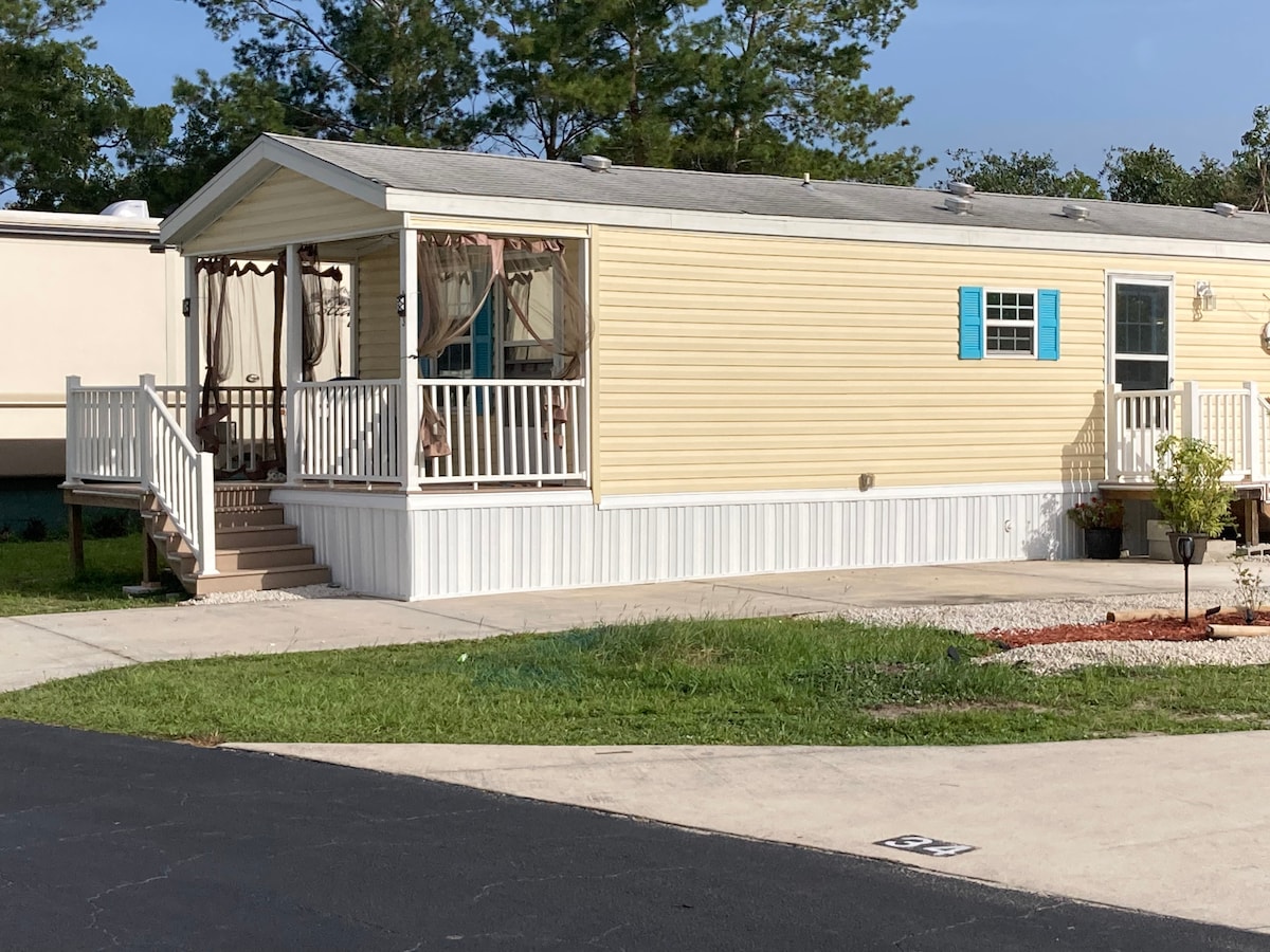 A charming yellow cottage with white trim is surrounded by a well-maintained landscape. A spacious front porch with railings invites relaxation, while decorative shutters add a pop of color. The cottage is positioned on a paved path, with lush green grass and decorative stones nearby.