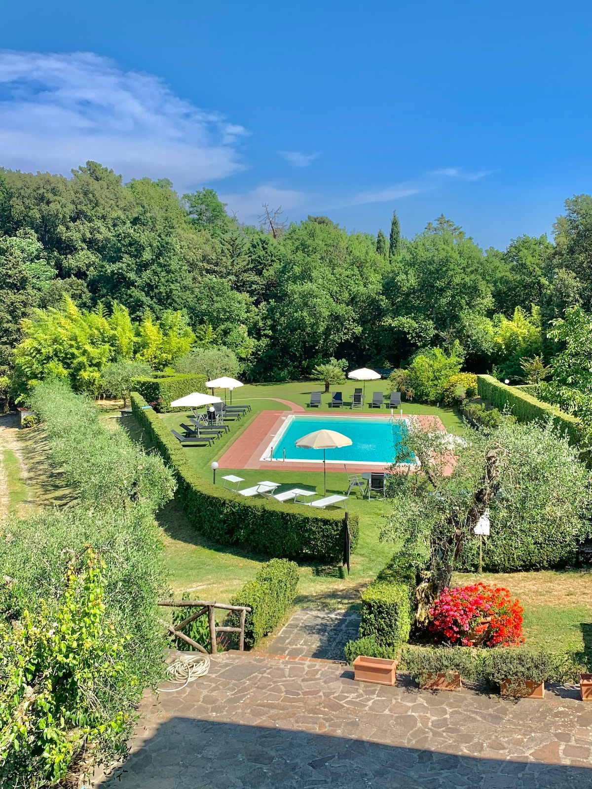 The outdoor area features a well-maintained garden surrounding a swimming pool, complemented by sun loungers and umbrellas. Lush greenery and colorful flowers create a serene atmosphere, with pathways leading through the beautifully landscaped grounds.