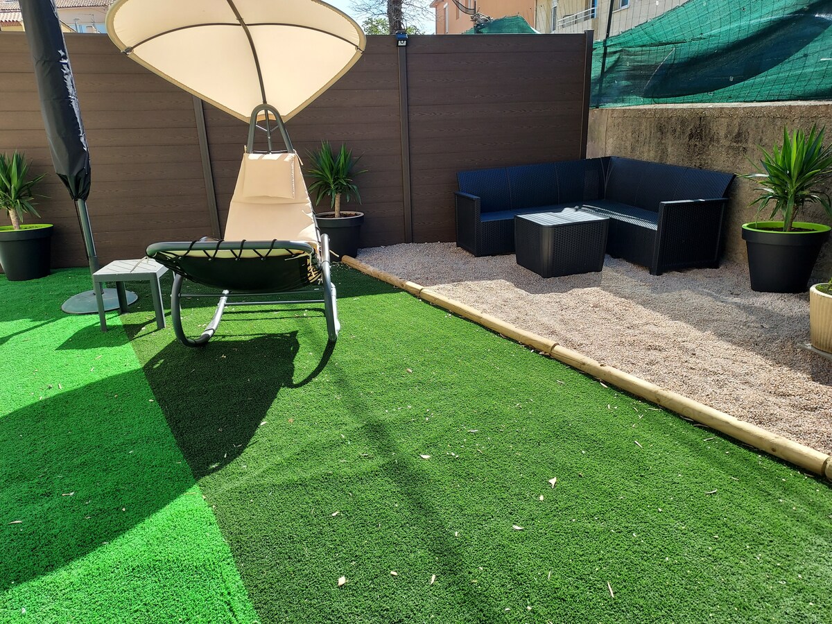 A private garden area features artificial grass and a sandy section. A comfortable seating arrangement consisting of a dark sectional sofa and a low table is positioned next to potted plants. A sun canopy shade provides comfort over a lounging swing chair.