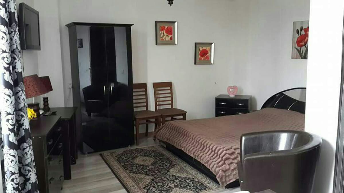 A sleeping area features a spacious bed covered with a beige blanket, framed by two decorative pieces of wall art. A black wardrobe and a dark dresser are present, complemented by two wooden chairs and a round table, enhancing the room's functionality.