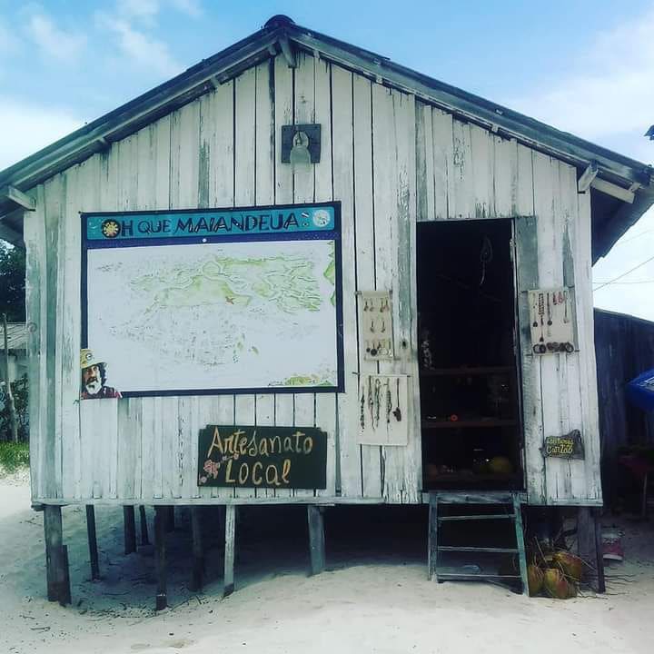 A rustic wooden structure stands on stilts, featuring a large map displayed on the front wall and various handmade crafts showcased around the entrance. The sandy ground surrounding the building enhances its beachside charm.