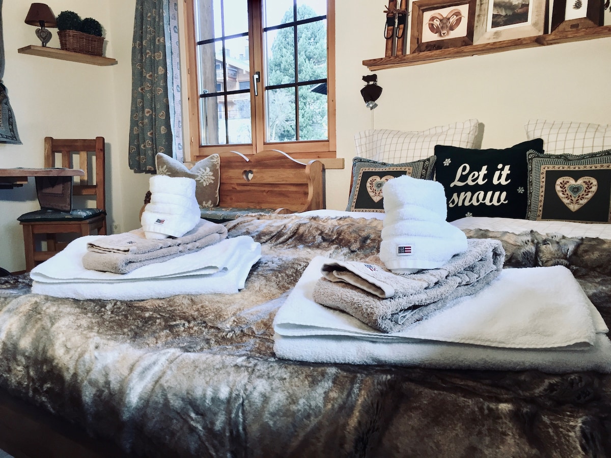 A cozy sleeping area features a spacious bed adorned with a warm, textured blanket. Soft towels are neatly arranged at the foot of the bed, while a decorative throw pillow adds a touch of charm. Natural light streams in through the wooden-framed window.