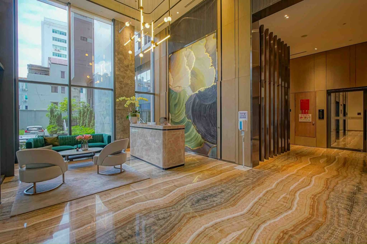 The lobby area features a modern reception desk surrounded by vibrant green plants. Comfortable seating is provided by two light-colored armchairs, positioned on a polished marble floor. A large artwork adorns the wall, creating an inviting atmosphere with natural light streaming in through large windows.