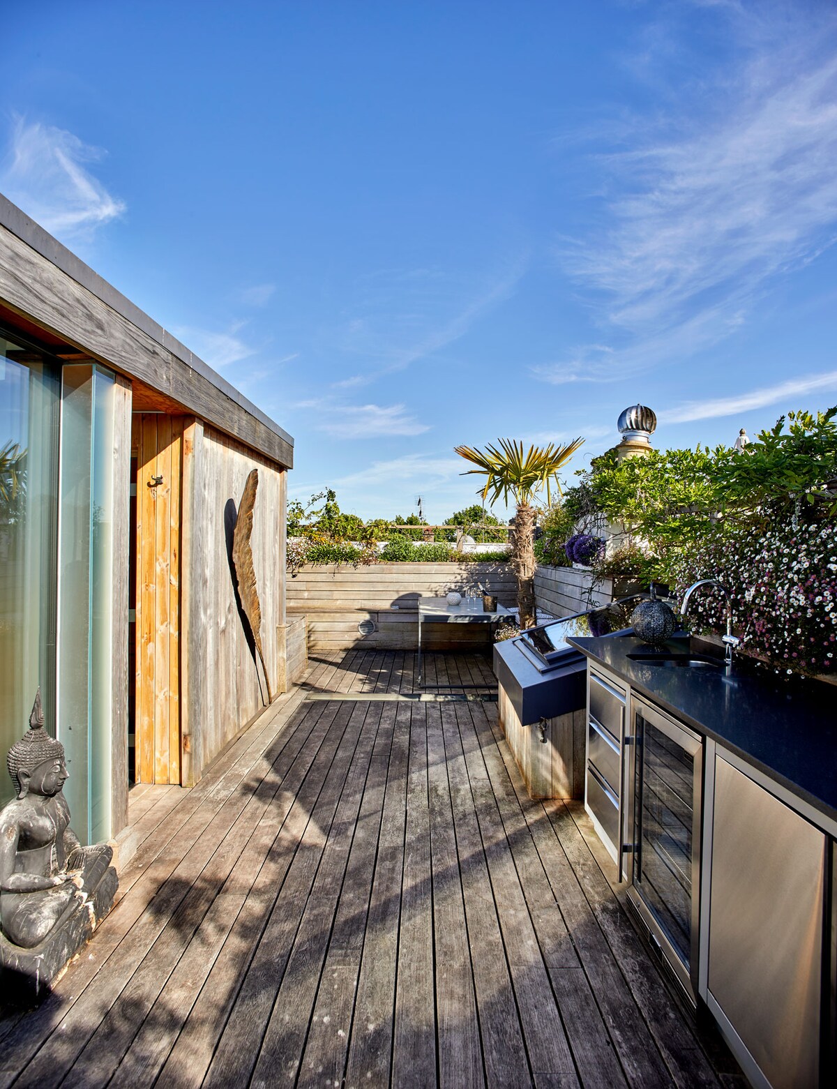 An outdoor roof terrace is showcased, featuring a wooden deck surrounded by greenery and decorative elements. A fully equipped outdoor kitchen with a sink and BBQ is visible, complemented by a serene statue. The scene is set against a clear blue sky.