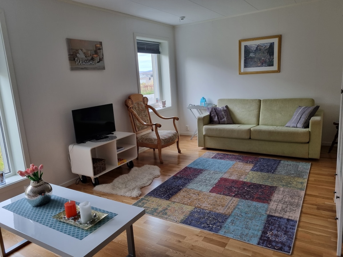 The living room features a soft green sofa positioned across from a small TV on a white stand. A cozy armchair is placed beside a window, while a patterned area rug adds warmth to the wooden flooring. A coffee table topped with candles and flowers completes the space.