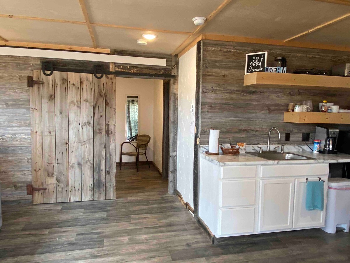 A kitchenette is showcased with white cabinetry and wood shelving, providing essential appliances and cookware. Natural wood accents create a rustic ambiance, with a sliding barn door leading to a cozy seating area. Soft light enhances the space, fostering a pleasant atmosphere.
