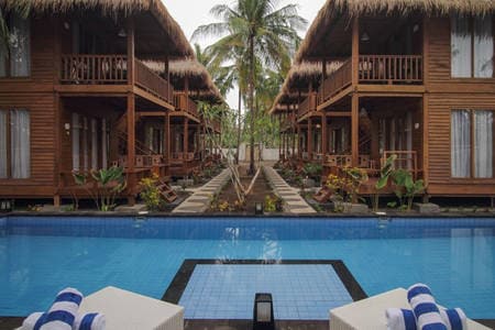The image captures a serene view of a swimming pool framed by traditional wood villas with thatched roofs. Lush greenery and tropical plants flank the pool area, while sun loungers are positioned at the edge, inviting relaxation in a tranquil setting.