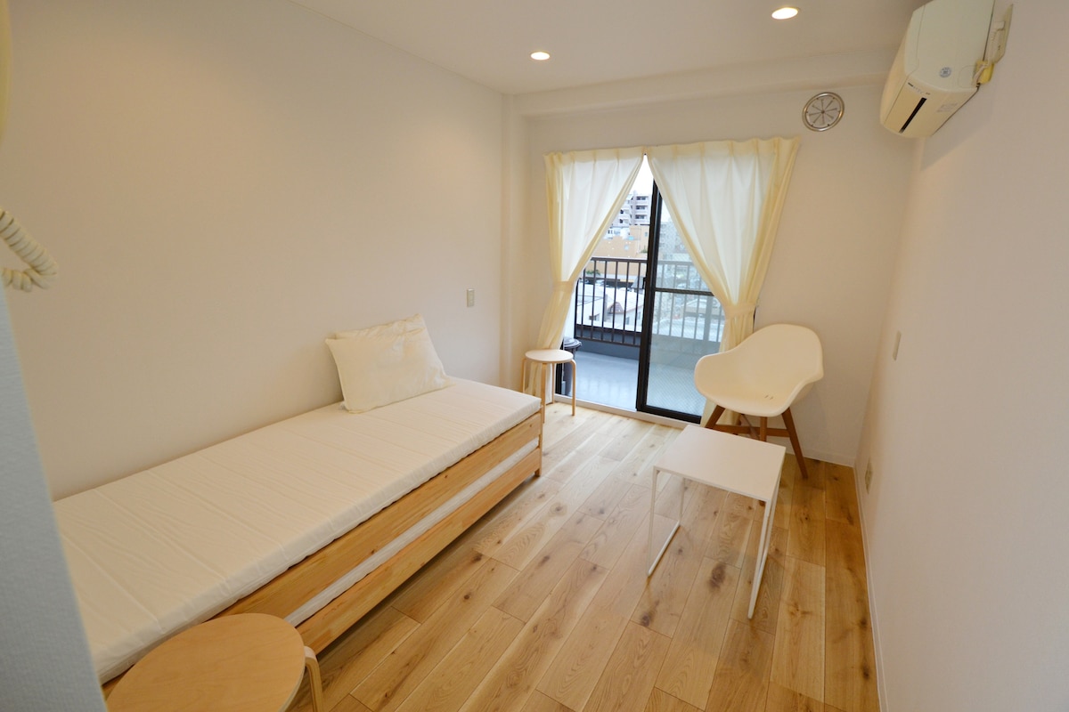 A bright room features a single bed with a white mattress and a soft pillow. A small round table and a minimalist chair are positioned beside a sliding glass door with sheer curtains, leading to a balcony. Light-colored wooden flooring enhances the spacious appearance.