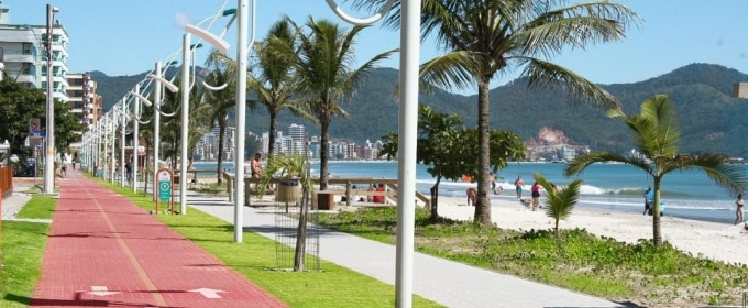 A vibrant beach promenade stretches alongside the shoreline, featuring a red bicycle path bordered by green grass and palm trees. Views of the ocean and distant hills can be seen, with people strolling along the sand and enjoying the coastal scenery.