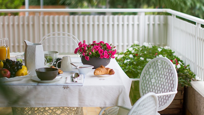 A terrace area is presented, featuring a white table set with dishes, cutlery, and a pitcher, alongside pastries and fruits. A container filled with vibrant pink flowers adds a touch of color, while white chairs surround the table, creating an inviting outdoor dining space.