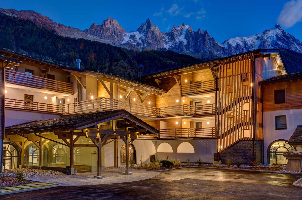 The exterior of the residence is illuminated at dusk, showcasing a blend of modern architecture and traditional materials. Wooden accents and warm lighting create an inviting entrance, framed by mountains in the background, featuring sharp peaks against a twilight sky.