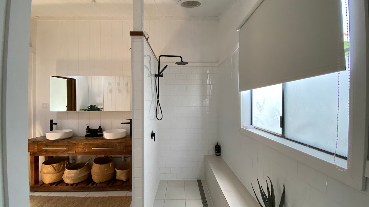 A generously sized bathroom is presented, featuring two vessel sinks mounted on a rustic wooden vanity. A spacious walk-in shower with a sleek black showerhead is visible, alongside a sunlit window with a roller shade. Natural elements are subtly incorporated with potted plants.