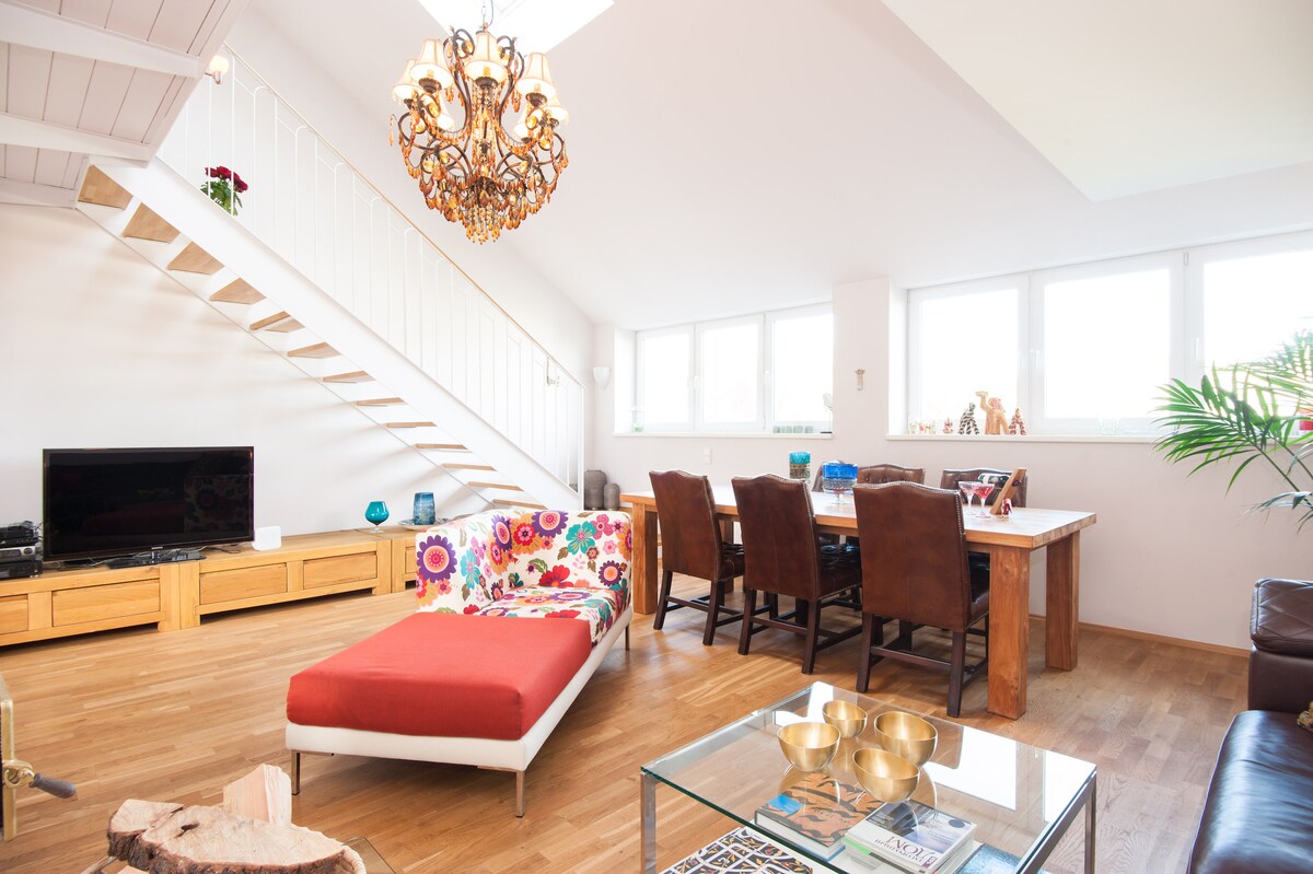 A spacious living area features a colorful patterned chaise lounge and a large wooden dining table surrounded by upholstered chairs. A flat-screen TV is positioned on a wooden console, and natural light fills the room through multiple windows. A staircase leads to the upper level.