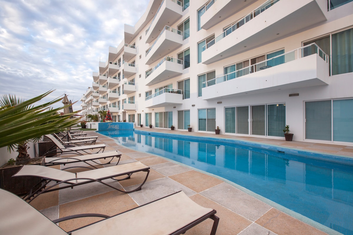 A long, narrow pool is framed by a row of lounge chairs, positioned along the tiled decking. Modern white buildings with multiple balconies stand in the background, while planters with greenery create a welcoming ambiance around the pool area.