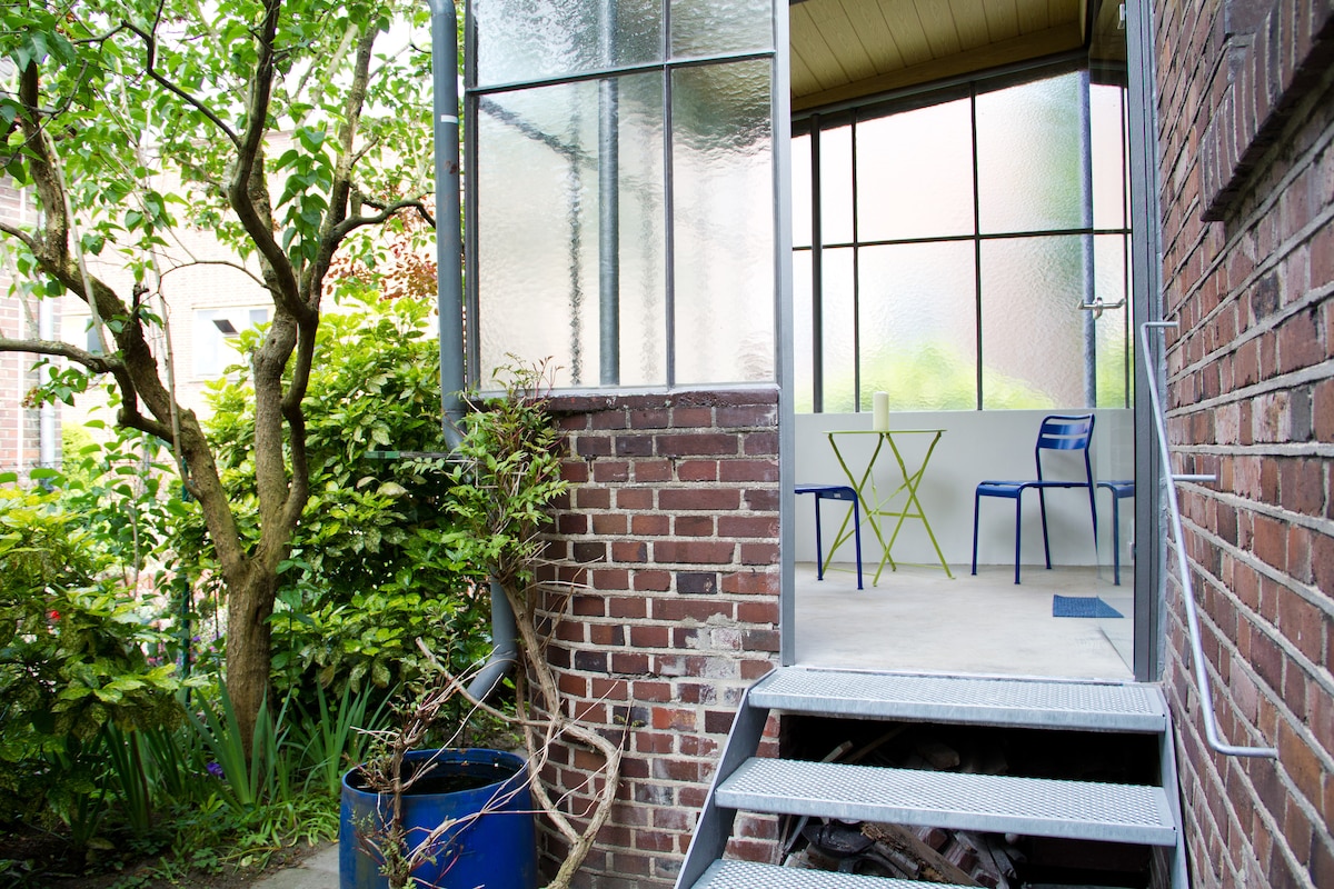 An entrance showcases a small set of stairs leading to a winter garden, designed with large glass panels allowing natural light to fill the space. Two chairs with a small table are positioned inside, surrounded by greenery and a brick wall backdrop.