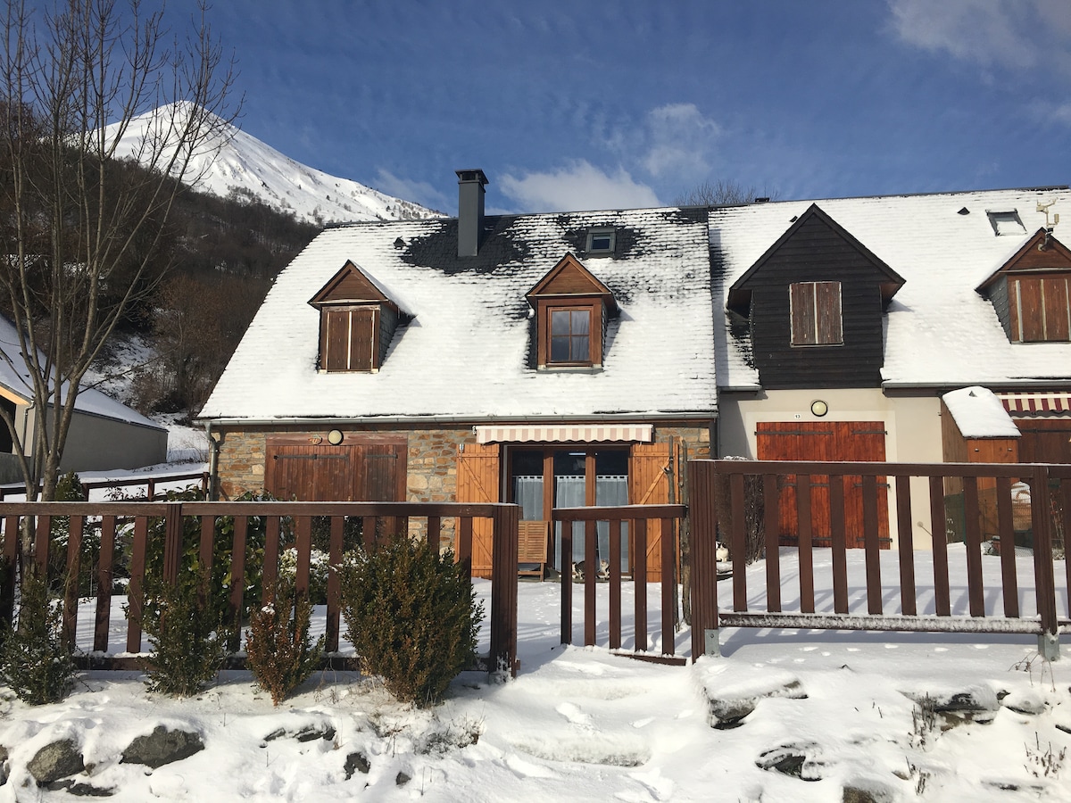 A charming duplex is surrounded by a snowy landscape, with wooden shutters adorning the windows. The exterior features stone accents, creating a rustic ambiance. A fenced area includes shrubs, while a mountain is visible in the background under a clear blue sky.