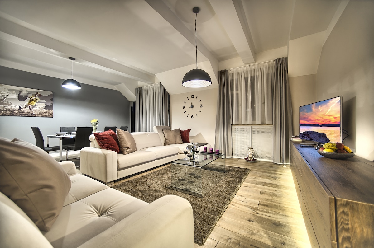 A modern living room features a seating area with a light-coloured couch adorned with pillows. A glass coffee table sits on a wooden floor, complemented by a wall with a clock and a television. Natural light filters through sheer, light-coloured curtains.