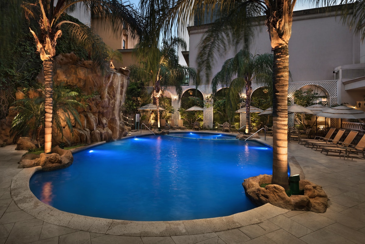 A large outdoor pool is surrounded by palm trees, with soft lighting enhancing its deep blue hue. A waterfall feature cascades over rocks, and comfortable lounge chairs are arranged along the poolside, creating a relaxing space for guests.