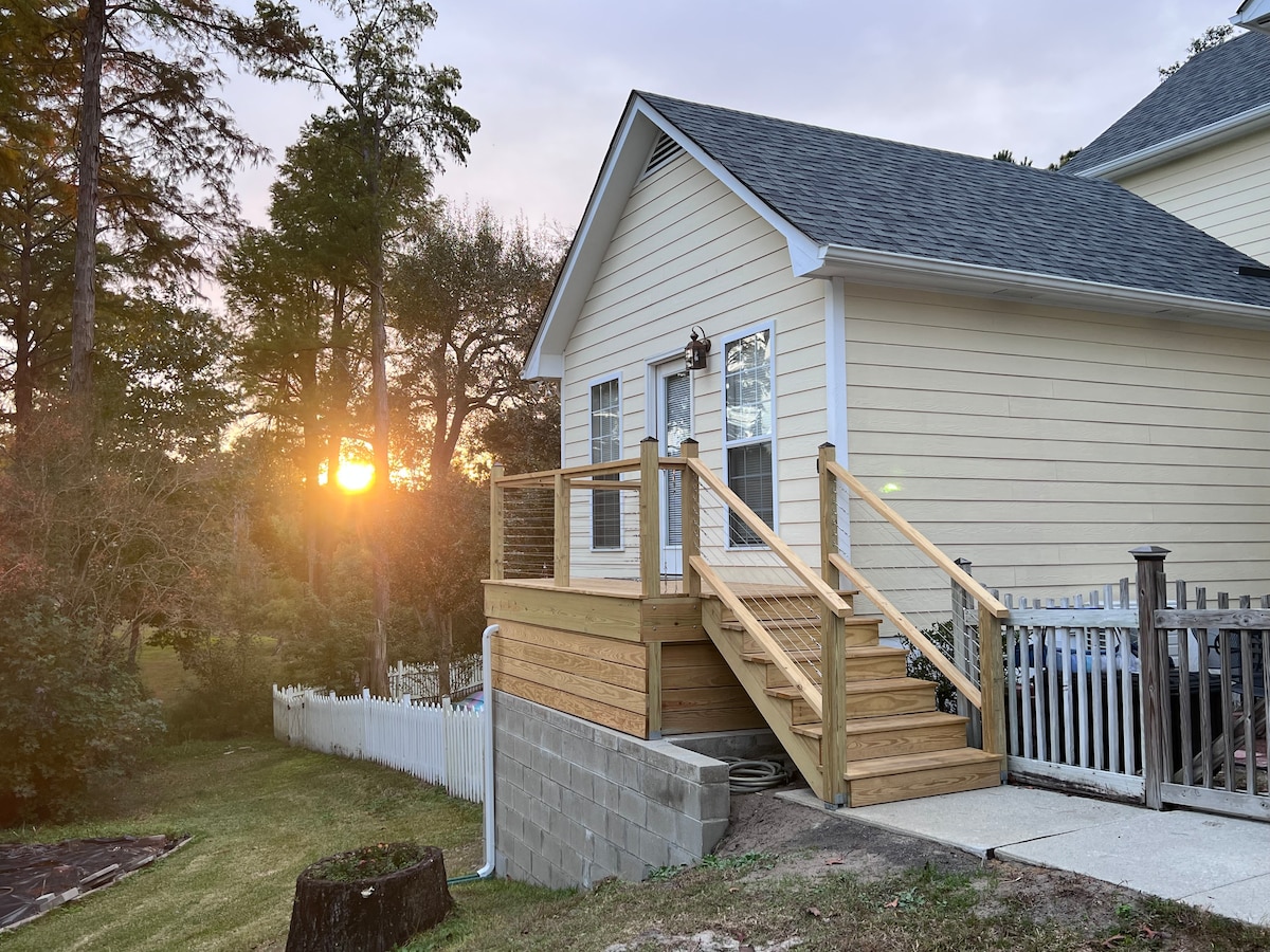 The exterior of a welcoming guest house is shown at sunrise, with warm light filtering through trees. A wooden deck with railings leads to the entrance, and a small pathway is visible along the side. A grassy area surrounds the home, adding to its peaceful setting.