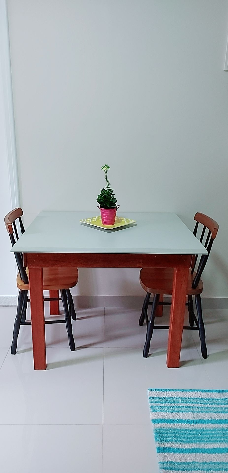 A dining table with a light gray surface is paired with two wooden chairs. At the center of the table, a small potted plant is placed atop a yellow tray. A striped rug in shades of blue lies on the floor beneath the table.