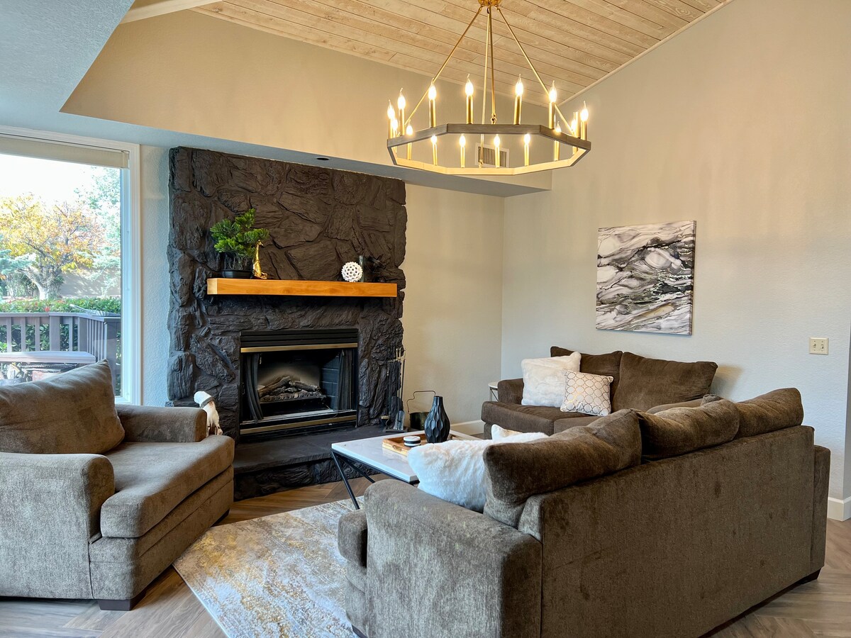 An inviting living area features a cozy seating arrangement with two plush sofas and a chair, set against a textured stone fireplace. A modern chandelier hangs above, illuminating the space. Natural light flows in through large glass doors, enhancing the bright atmosphere.
