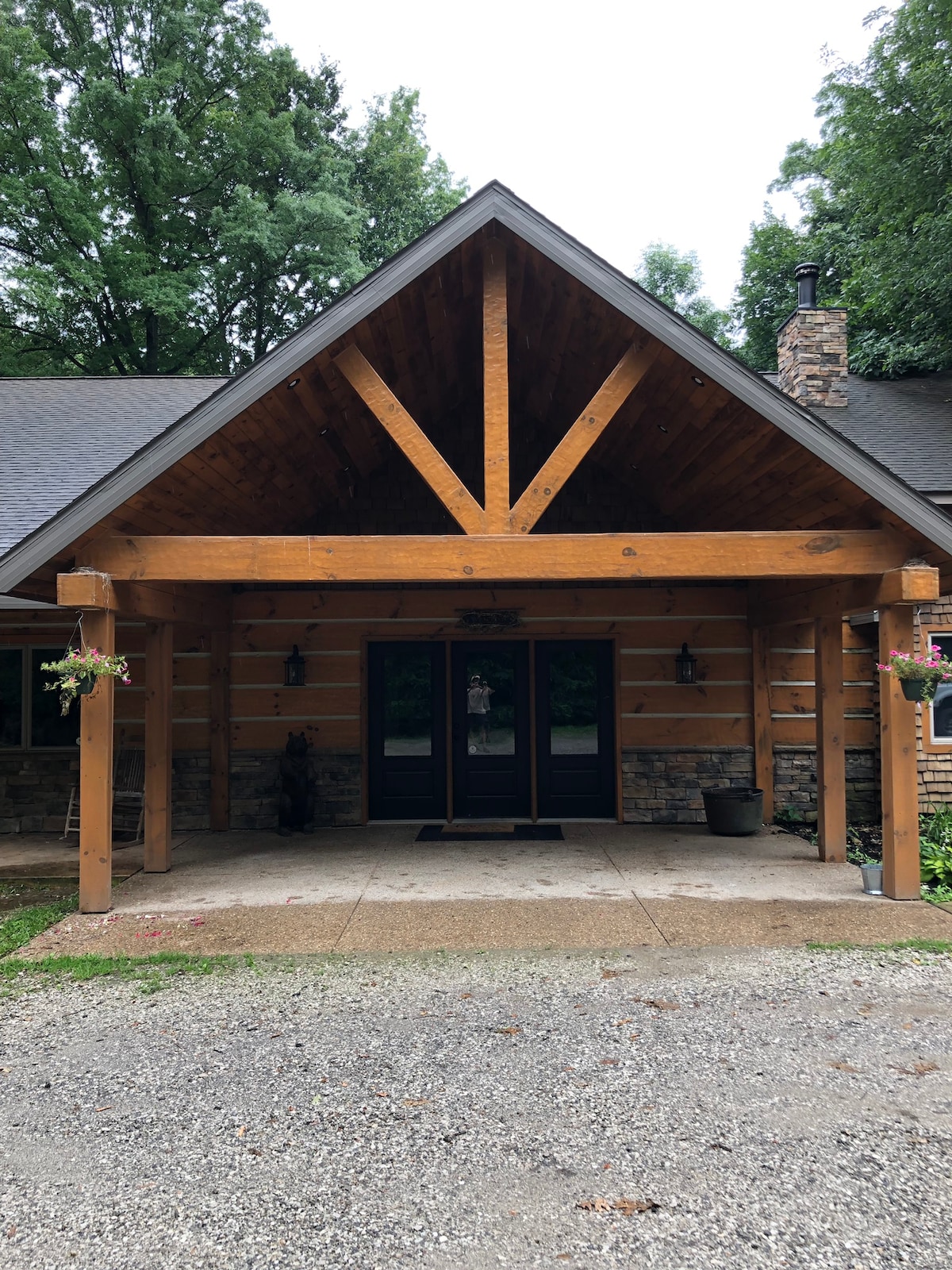 The entrance features a striking wooden overhang supported by large beams, creating a welcoming shelter. A stone accent wall complements the lodge's rustic design. The double front doors provide an inviting entry point, flanked by decorative wall sconces and hanging floral arrangements.