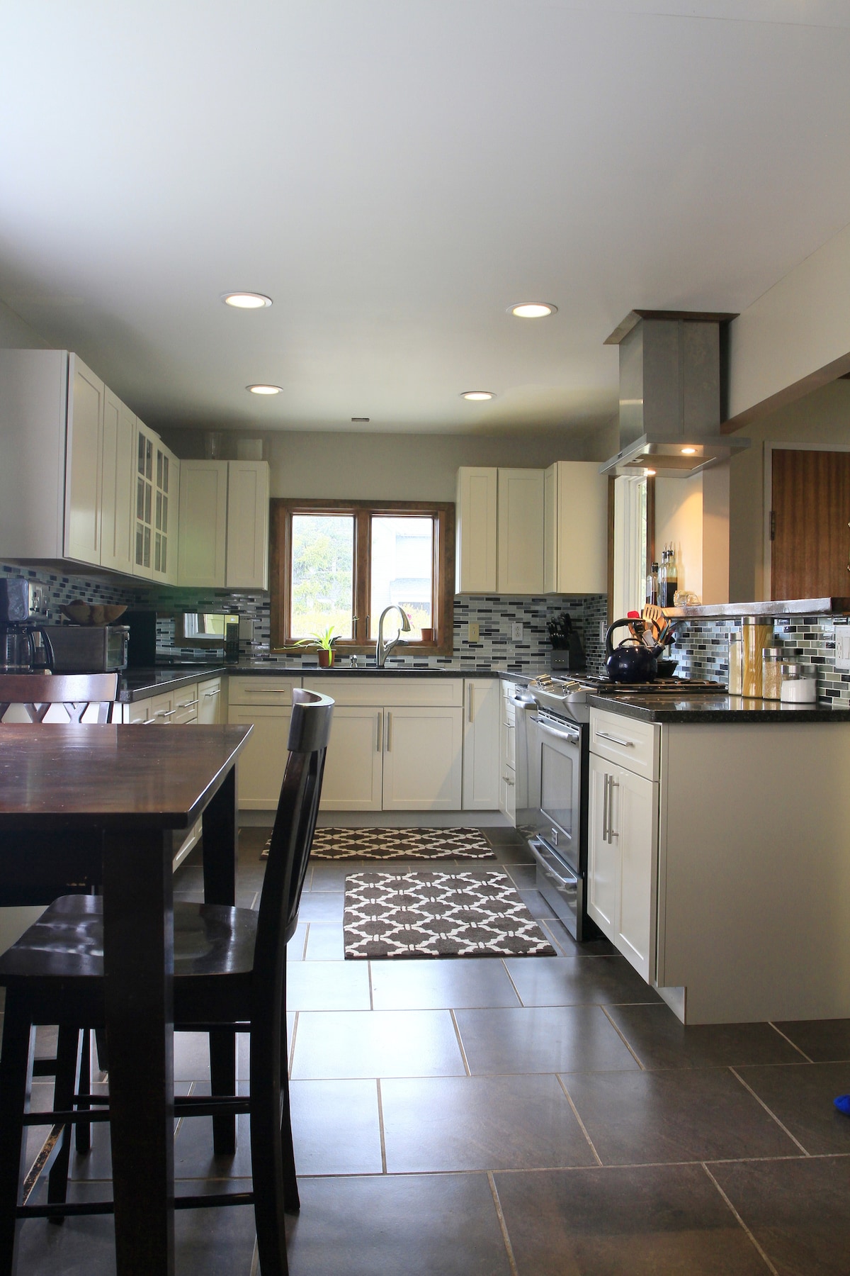 A modern kitchen features sleek cabinetry and stainless steel appliances. Natural light enters through a window above the sink, illuminating the dark countertops and tile flooring. A dining table with high chairs is visible, accompanied by a decorative rug that adds warmth to the space.