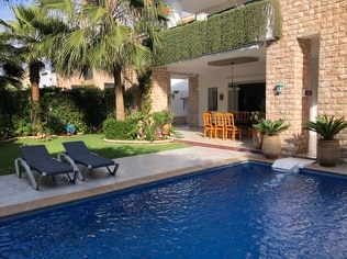 A private outdoor area features a clear blue swimming pool bordered by a stone deck. Two lounge chairs are positioned beside the pool. Lush greenery surrounds the space, complemented by a dining area set with chairs in the background, creating a serene and inviting environment.