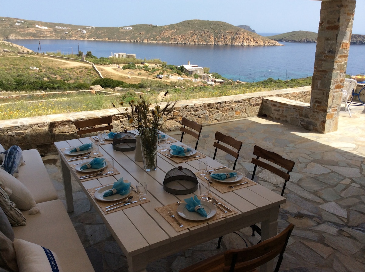 An outdoor dining table is set for a meal, featuring plates, utensils, and decorative blue accents. Surrounding the stone patio, the view includes a tranquil sea and hills in the distance. The setting offers an inviting space for group meals while enjoying the scenery.