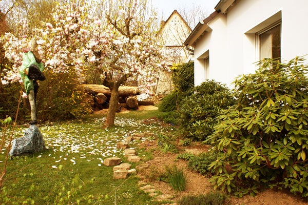 A well-maintained garden is shown, featuring a blossoming tree with pink flowers beside an artistic sculpture. Neatly arranged stones form a path through the greenery, while logs are visible in the background, enhancing the natural setting.