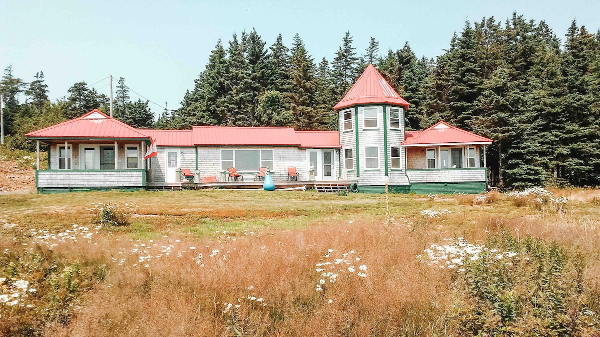 The exterior of a unique cottage is showcased, featuring a turret and a red roof. Surrounding greenery is complemented by wildflowers across an open yard. A patio area with chairs offers a comfortable space to enjoy the views of nature.