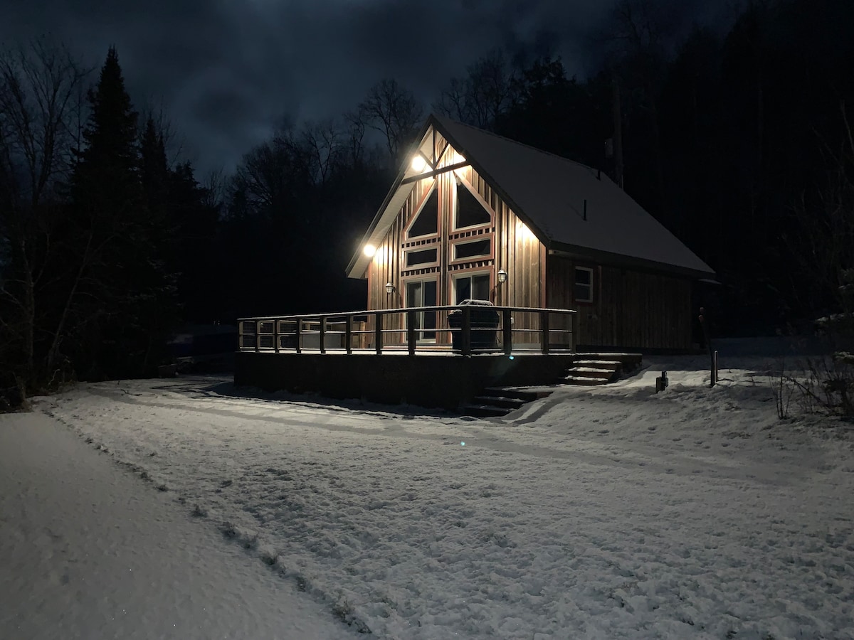 A cottage is showcased at night, featuring wooden exterior walls and an angled roof. Illuminated by lights, the front porch is visible, and snow blankets the ground, creating a serene winter landscape. Trees are faintly seen in the background under a cloudy sky.