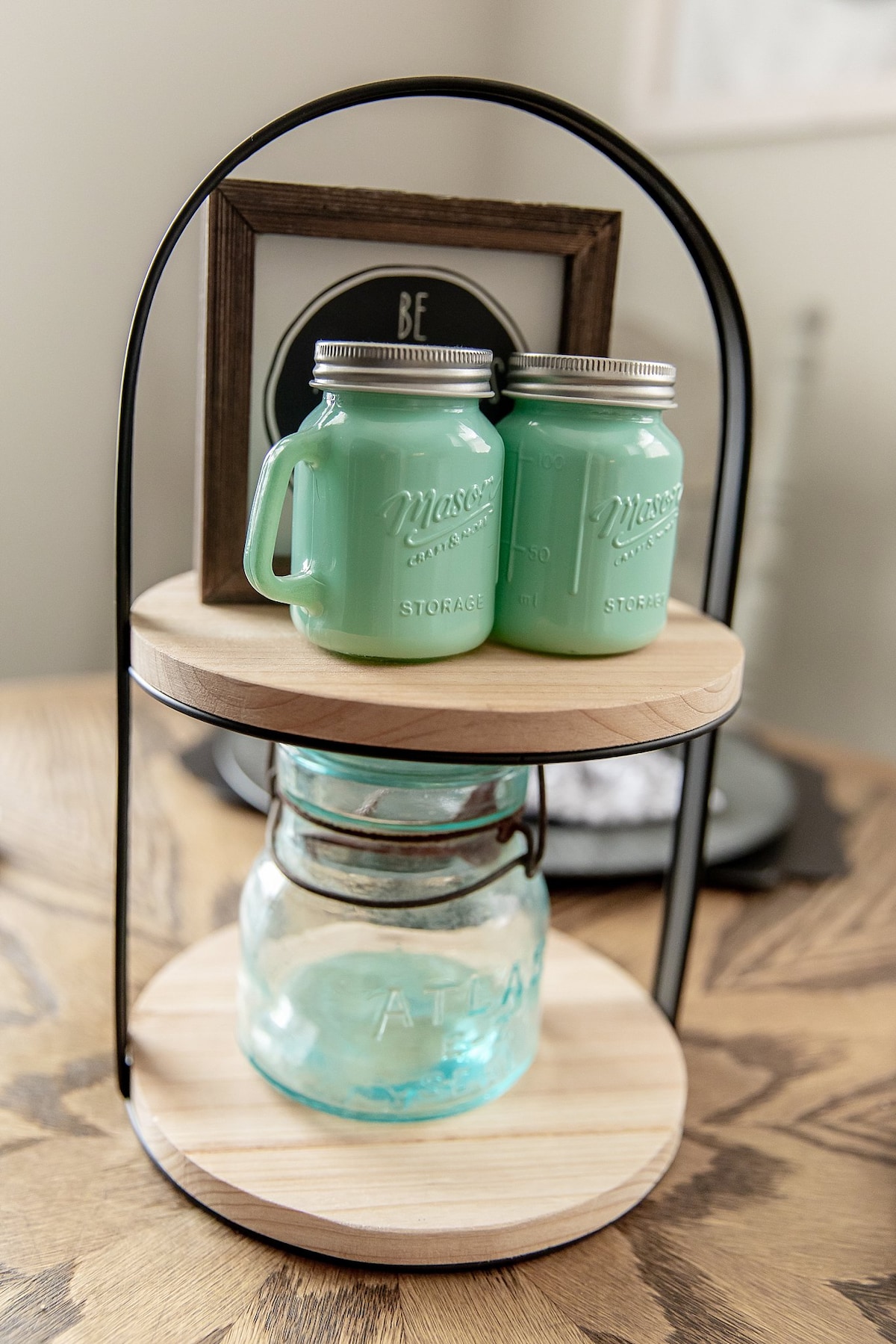 A two-tiered storage display features mason jars in pastel green, complemented by a glass jar on the lower level. A framed decorative piece sits on top, adding a personal touch to the wooden display. The setup is placed on a textured wooden surface.