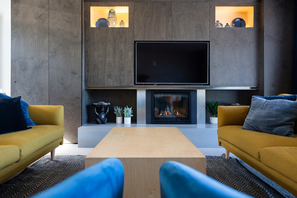 A modern living area features a sleek wall-mounted television above a gas fireplace, set against textured wood paneling. Two comfortable sofas in vibrant colors flank a minimalist coffee table, while decorative items and plants add subtle accents to the space.