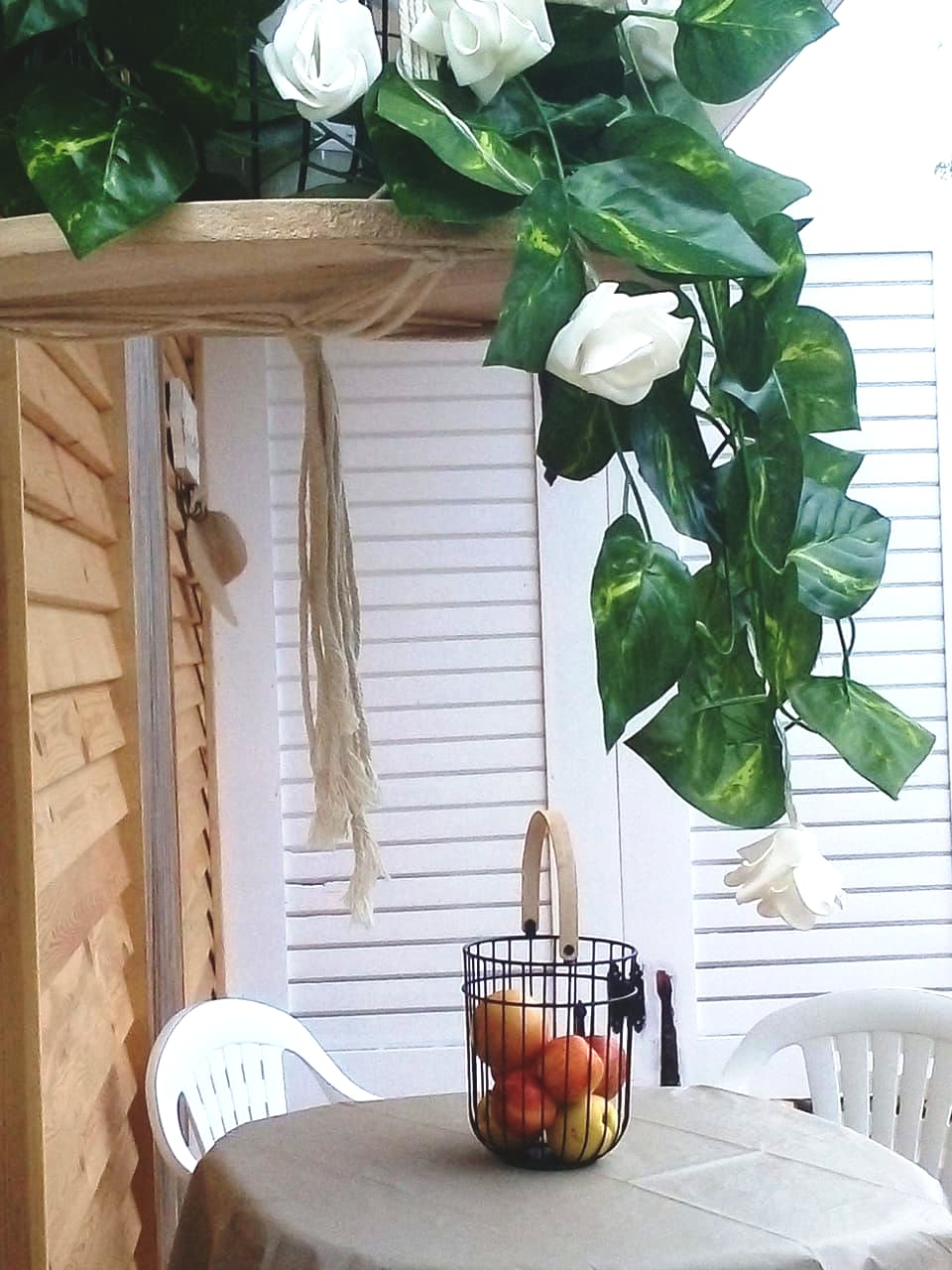 A table covered with a neutral fabric is seen, accompanied by two white chairs. A wire basket filled with colorful fruits sits in the center, while artificial flowers and greenery hang above, adding a touch of color to the outdoor setting. Shuttered doors are visible in the background.