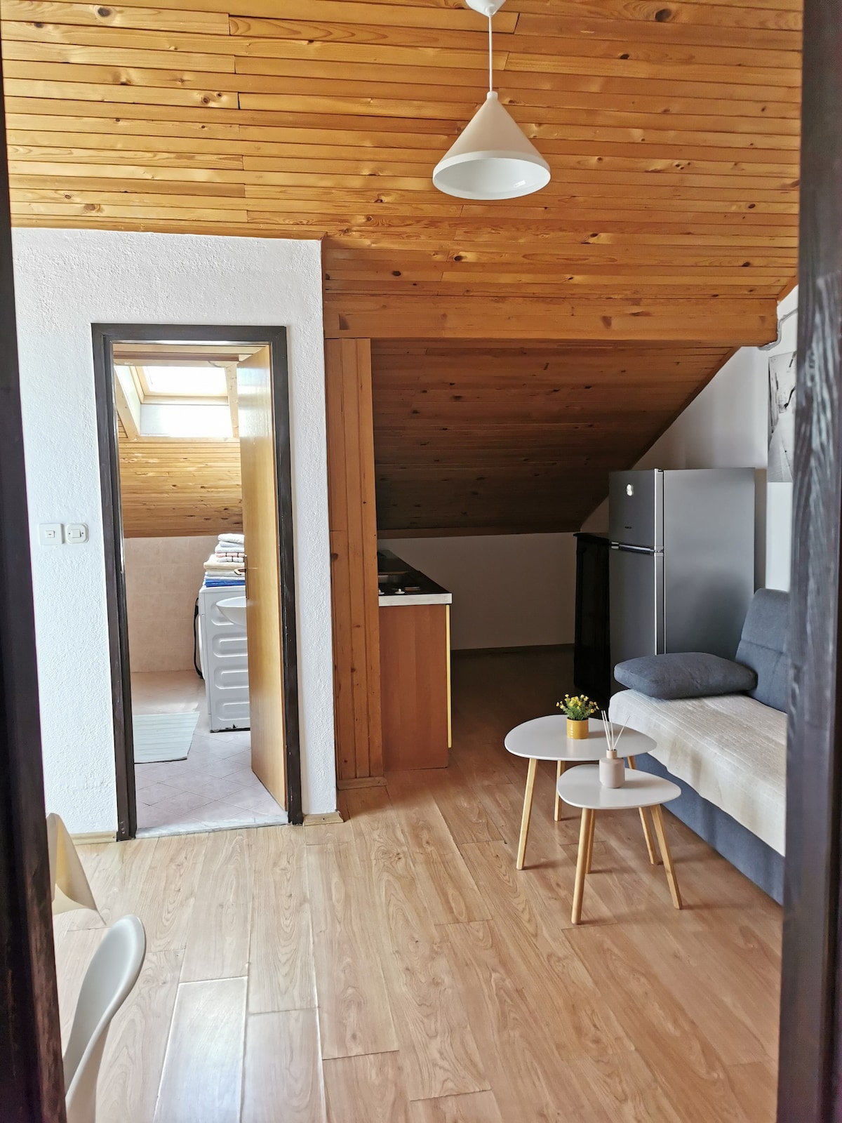 A loft space is captured from an entryway perspective, highlighting the warm wooden ceiling and light-colored walls. Two small tables are positioned in front of a sofa, with a kitchen area visible in the background. A window allows natural light to brighten the room.