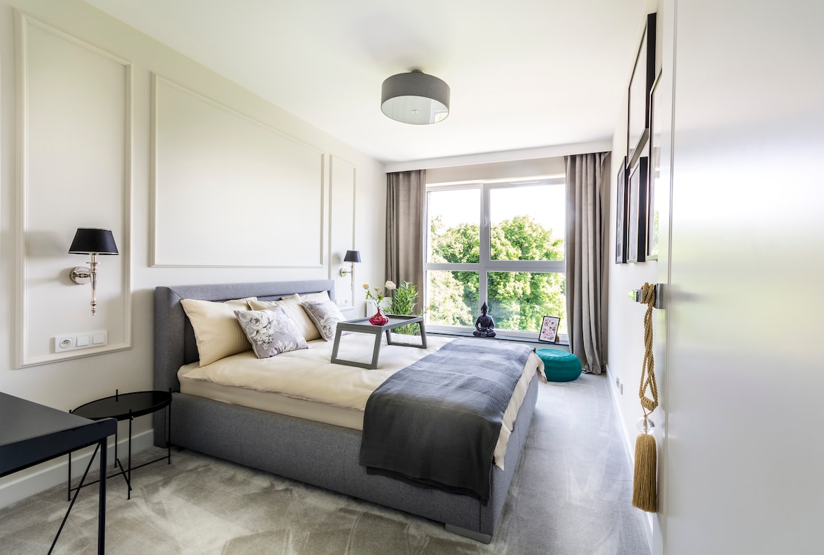 A calming bedroom is furnished with a spacious bed, accented by neutral textiles. Natural light filters through large windows, framing a view of greenery outside. A small side table and decorative elements add character, while soft shades create a serene atmosphere.