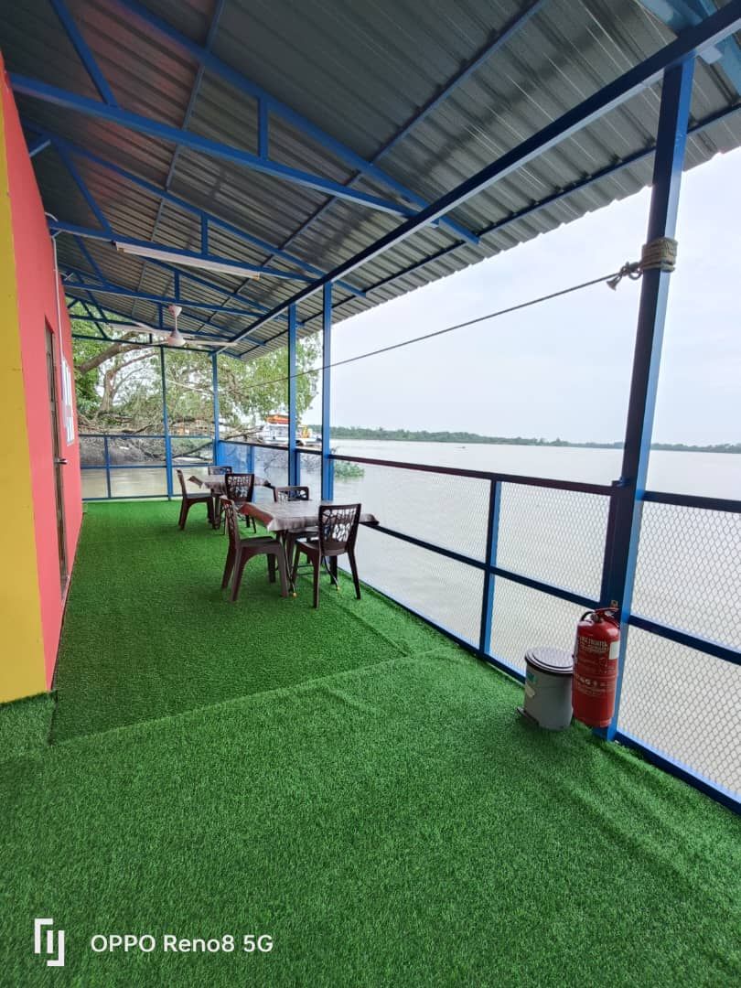 An outdoor seating area is presented with several tables and chairs arranged on a green artificial turf surface. A covered deck overlooks the river, with safety features observed nearby, enhancing the serene riverside experience.